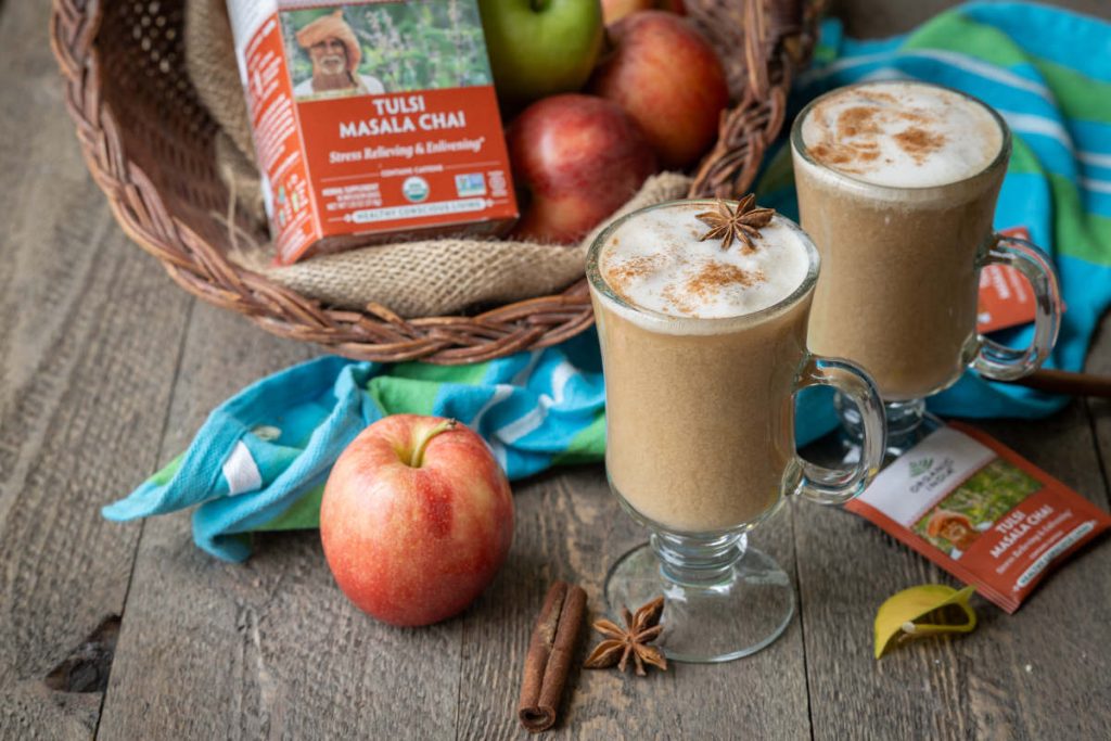 Two mugs of apple chai latte on a picnic table with basket of apples.