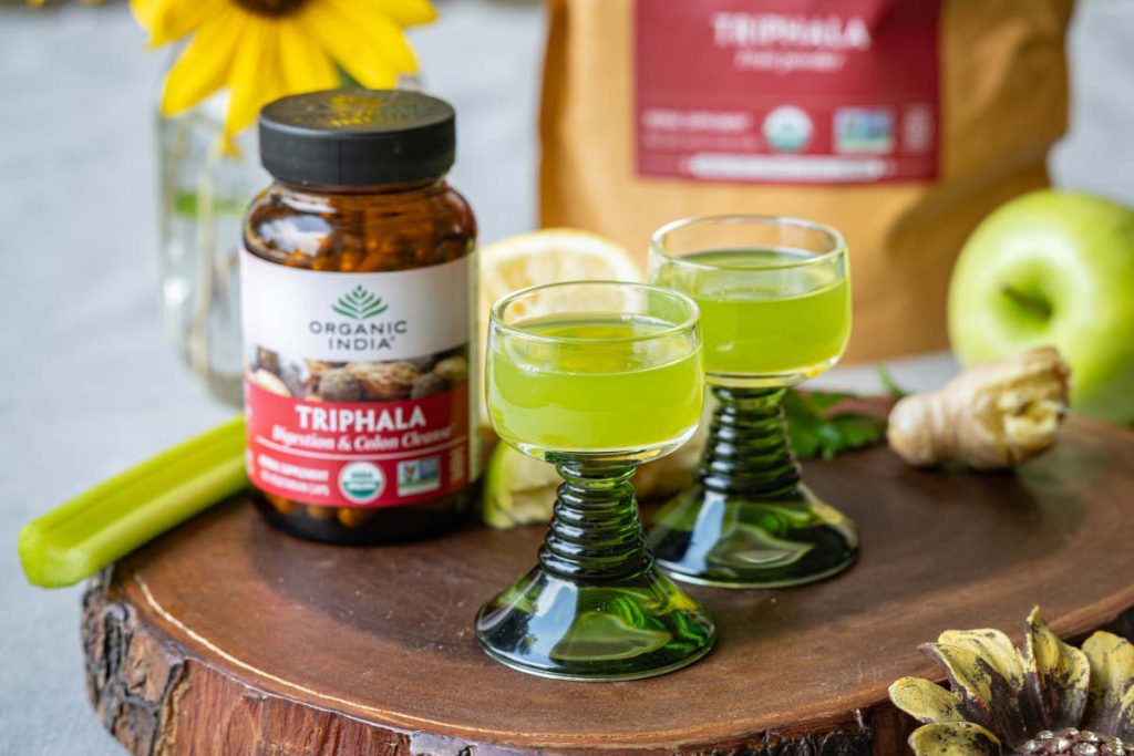 Triphala apple detox shot with celery and apple on a wood cutting board.