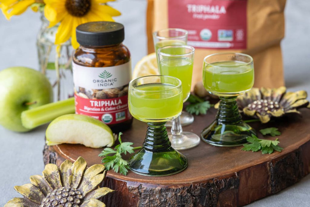 Triphala apple detox shot with celery and apple on a wood cutting board.