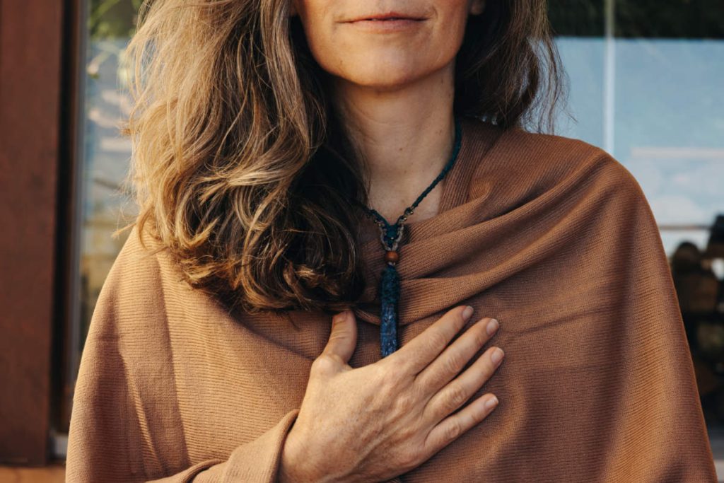 Ayurveda for women wearing a brown poncho with hand at her heart and handmade necklace.