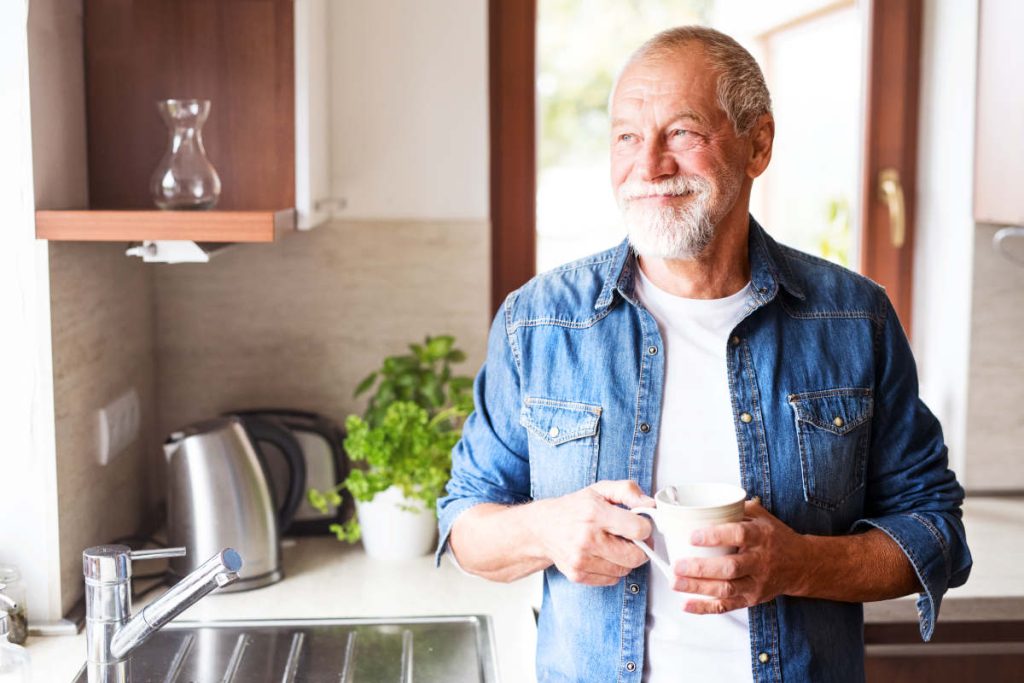 Older man in kitchen with herbs and plants drinking a cup of tea.