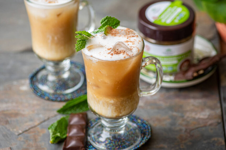 Iced chocolate moringa latte with micro-foam in glass with chocolate and moringa leaves scattered on rustic tile table.