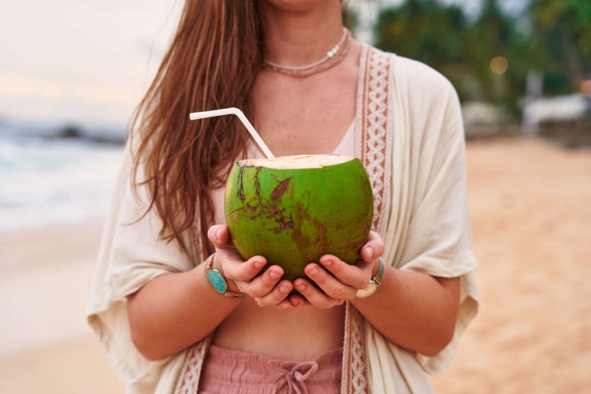 Woman drinking from coconut on beach for her late summer ayurveda ritual