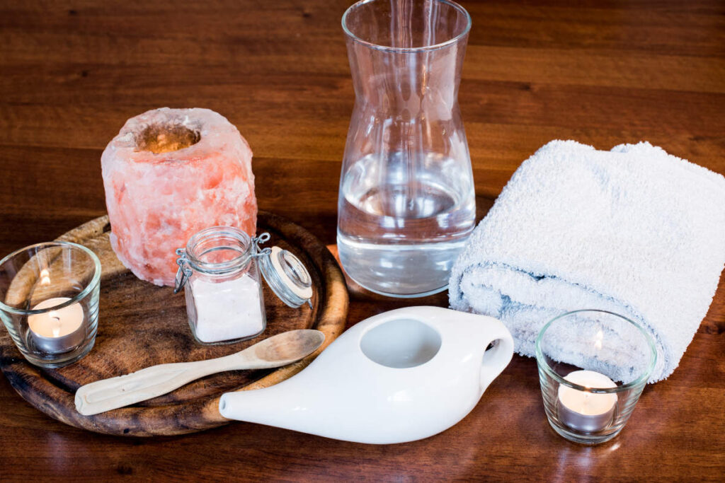 Neti pot for spring sensitivities on a wooden table with candle, hand towel, water, and Himalayan pink salt.
