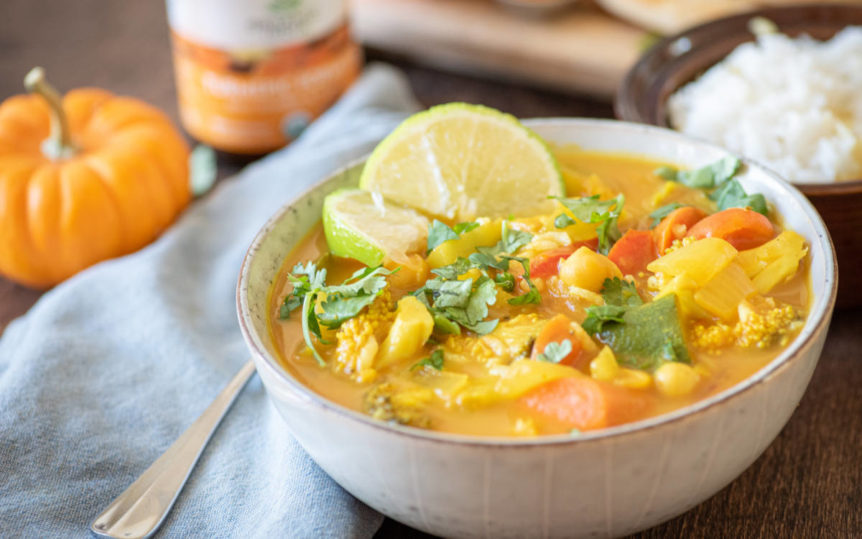 A white bowl full of pumpkin curry with a pumpkin, turmeric supplement and rice behind it.