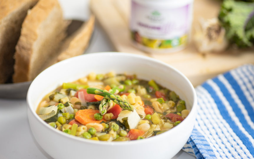 Spring minestrone in a white bowl on a table nearby a blue and white dish towel.