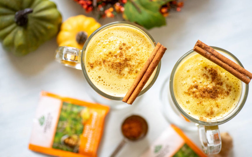 Two clear mugs to the brim with frothy pumpkin turmeric latte with cinnamon sticks on white table.